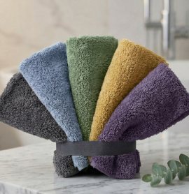 A neatly stacked set of four folded EXLUX premium skincare facial towels in yellow, green, purple, and stone grey on a warm wooden surface
